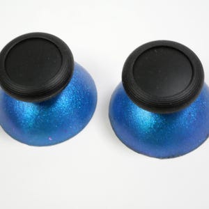 Może przedstawiać: Dwa ozdobne korki do butelek. Każdy korek ma czarną, okrągłą górę z żebrowanym brzegiem, osadzoną na niebieskiej, iryzującej, zaokrąglonej podstawie. Niebieska podstawa ma połyskujący, błyszczący efekt.