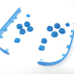 May include: Blue, glitter-flecked plastic components, including curved frame sections and various round and square buttons, are displayed. The parts are arranged on a white surface, possibly from a disassembled electronic device or a craft project.