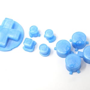 May include: A selection of light blue game controller buttons, including a D-pad, square, and round buttons. The buttons are made of a solid material and are arranged on a white surface.