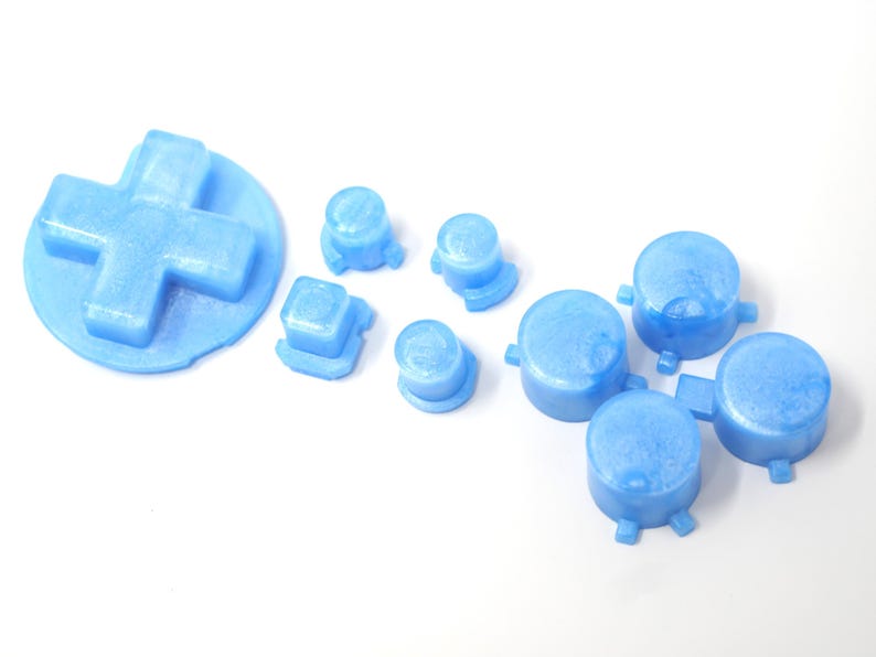 May include: A selection of light blue game controller buttons, including a D-pad, square, and round buttons. The buttons are made of a solid material and are arranged on a white surface.