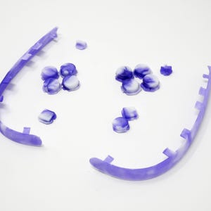 May include: Two curved, lavender-colored plastic pieces with a series of rectangular notches. Scattered around are small, round, purple and white marbled pieces. The items are on a white background.