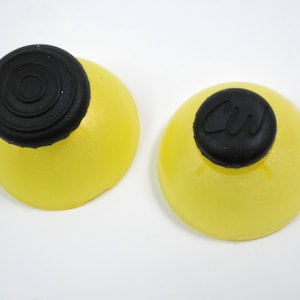 Puede incluir: Dos objetos redondos amarillos con tapas negras. Una tapa tiene círculos concéntricos, la otra tiene las letras "CW". Los objetos están sobre un fondo blanco.