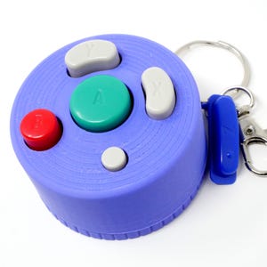 May include: A purple keychain shaped like a video game controller. It has a red button, a green button with the letter "A", and two gray buttons. The keychain includes a blue tag and a silver keyring and clasp.