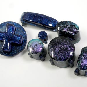 Könnte beinhalten: Eine Sammlung von Gaming-Zubehör im Galaxy-Design. Das Set beinhaltet ein Steuerkreuz, Knöpfe und eine lange Leiste, alle mit einem dunkelblauen und violetten Glitzereffekt, der einen kosmischen Look erzeugt.