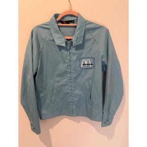 Puede incluir: Una chaqueta azul claro con cremallera, cuello clásico, mangas largas con puños abotonados y dos bolsillos delanteros. Un parche rectangular en el pecho izquierdo muestra el logotipo blanco de McDonald's con el nombre en azul oscuro.
