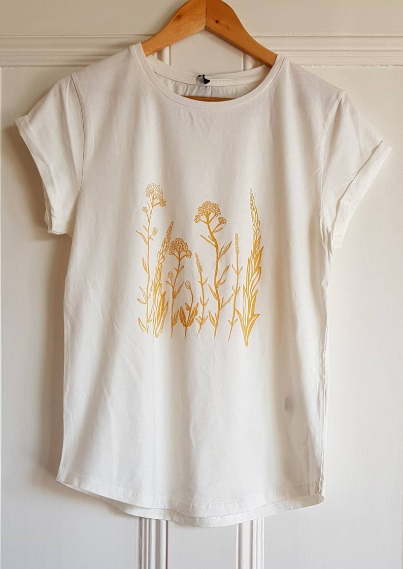 May include: White t-shirt with a yellow floral design. The design features a cluster of stylized flowers and leaves.