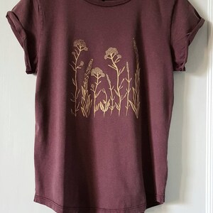 May include: A maroon t-shirt with a floral design printed in a light yellow color.