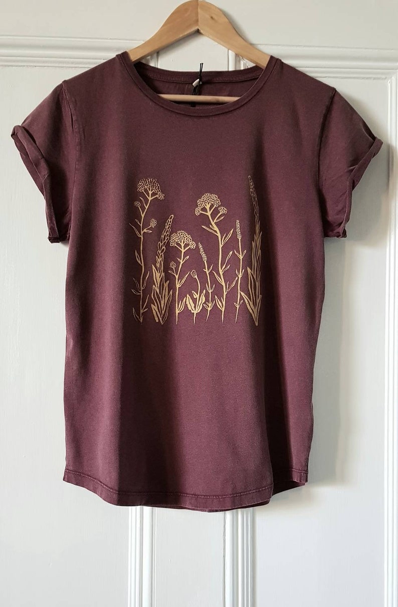 May include: A maroon t-shirt with a floral design printed in a light yellow color.