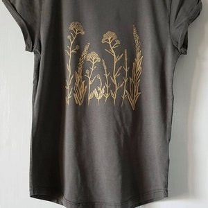 May include: A dark gray t-shirt with a gold floral print. The print features a variety of stylized flowers and leaves.