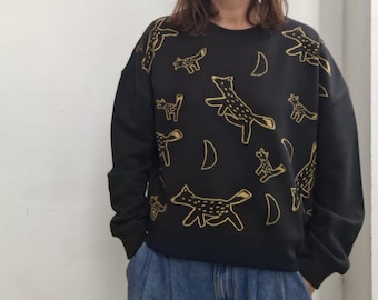 May include: Black sweatshirt with a repeating pattern of gold-colored foxes and crescent moons. The casual top has a crew neck and long sleeves. The design is embroidered on the fabric. The sweatshirt is worn with blue jeans.