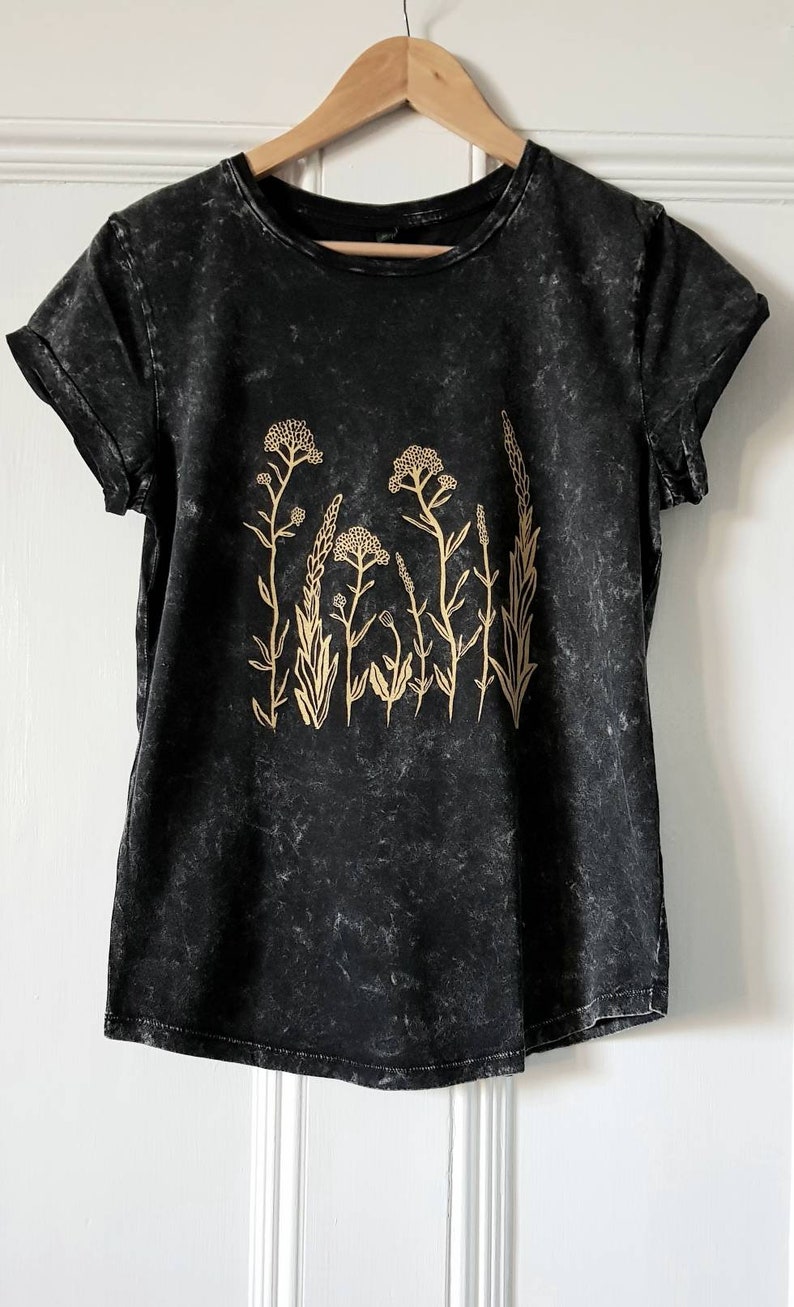 May include: A black t-shirt with a distressed look features a gold floral design. The design includes several stylized flowers and leaves.