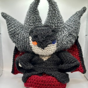 May include: A crocheted plush toy in the shape of a vampire with gray, black, and red details. The toy has large ears, a black cape, and a red dress.