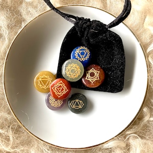 May include: A collection of seven chakra stones in different colours, each with a gold symbol, presented on a white plate with a gold rim. A black velvet drawstring bag contains some of the stones. The stones are for meditation and spiritual practices.