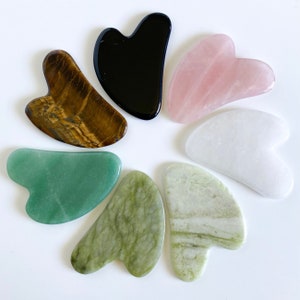 May include: A selection of seven gua sha tools in different colours and materials. These include tiger's eye, black obsidian, rose quartz, white, green, and two shades of green jade. Each tool has a curved, heart-like shape.
