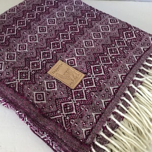 May include: A woven alpaca blanket with a geometric diamond pattern in shades of burgundy and white. The blanket has a fringed edge and a tag that reads "Tejarte Alpaca".