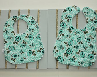 May include: Two turquoise and white floral print bibs with a scalloped edge. The bibs are hanging on a gray wooden board with twine.