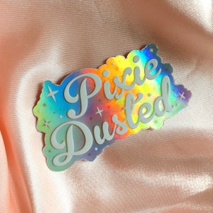 Puede incluir: Una pegatina holográfica con fondo arcoíris y estrellas blancas. La pegatina dice "Pixie Dusted" en una fuente blanca cursiva.