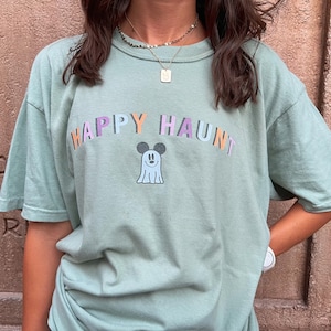 Puede incluir: Una camiseta verde claro con el texto "HAPPY HAUNT" en colores arcoíris. Un fantasma de dibujos animados con orejas de Mickey Mouse está en el centro del texto.