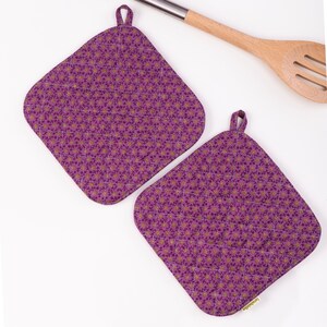 May include: Two quilted purple pot holders with a floral pattern and hanging loops. The pot holders are diamond-shaped with a quilted design. A wooden spoon is visible in the background.