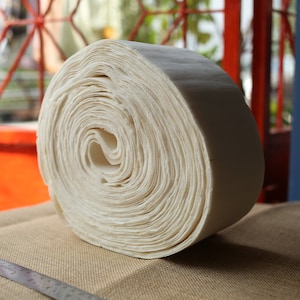 Puede incluir: Un tejido blanco roto enrollado apretadamente, que se asemeja a un rollo grande y grueso de papel, se encuentra sobre una superficie de arpillera marrón.