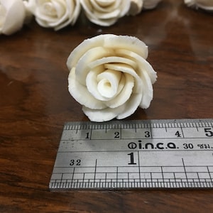 May include: A close-up of a single white wooden rose measuring 1 inch in diameter. The rose is sitting on a wooden surface next to a ruler.