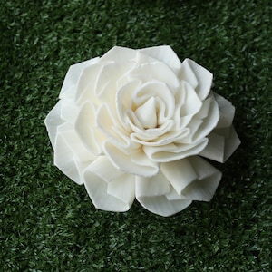 May include: A white, fabric flower with a ruffled design. The flower is on a green, artificial grass background.