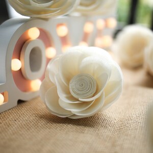 Puede incluir: Letras de madera blancas que deletrean "LOVE" con bombillas, decoradas con flores de papel blanco. Las flores están dispuestas sobre una superficie de arpillera.