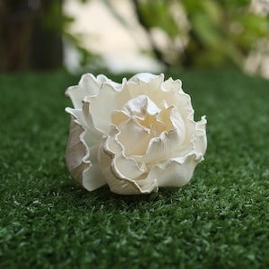 May include: A white, paper flower with ruffled petals, resembling a peony, is displayed on a green artificial grass surface.