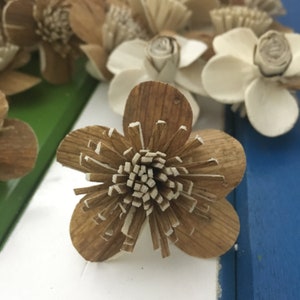 May include: A brown wooden flower with a white center. The flower has a layered design and is made of thin strips of wood.