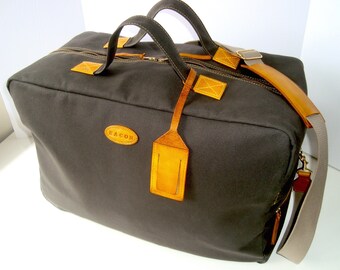 Pode incluir: Uma bolsa de viagem de lona cinza escura com detalhes em couro marrom e uma etiqueta de couro marrom. A bolsa tem fecho de correr e alça superior. A bolsa tem o nome da marca "MACON" na frente.