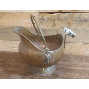May include: A polished brass coal scuttle featuring a wide pouring spout, a sturdy arched handle, and a pedestal base. The vessel is adorned with a decorative lion head mount at the handle attachment point and includes a secondary handle with a white ceramic grip featuring a blue floral pattern.