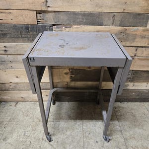 May include: A gray metal table with drop-leaf sides and a rectangular top. The table has a wheeled base and a distressed finish, suggesting it is vintage or has been well-used. The table is set against a wooden plank background.