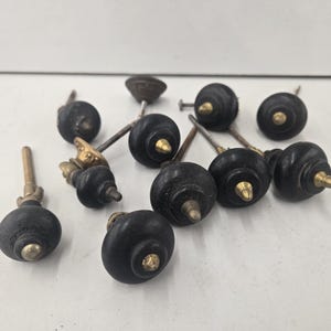 May include: A collection of nine drawer knobs featuring round black wooden heads with brass-toned metal centers and attached mounting screws. Each knob has a tiered, circular design with a central metallic point, arranged in a cluster.