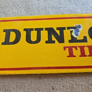 K&ouml;nnte beinhalten: Ein gelbes rechteckiges Schild mit dem Wort "DUNLOP" in gro&szlig;en schwarzen Buchstaben und "TIRES" in Rot. Das Schild hat einen roten Rand und ist leicht besch&auml;digt.