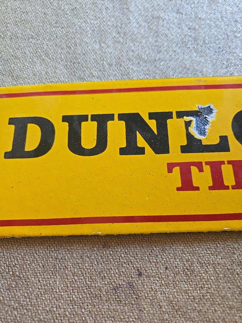 K&ouml;nnte beinhalten: Ein gelbes rechteckiges Schild mit dem Wort "DUNLOP" in gro&szlig;en schwarzen Buchstaben und "TIRES" in Rot. Das Schild hat einen roten Rand und ist leicht besch&auml;digt.