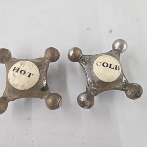 May include: Two vintage cross-shaped faucet handles with a metallic finish and worn patina. Each handle features a central white circular ceramic insert with black text, one labeled HOT and the other labeled COLD, with four rounded knobs.