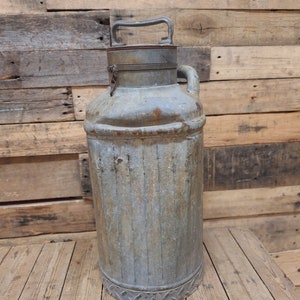 May include: A vintage metal milk can with a hinged lid and a handle. The can is gray and has a worn, rustic look. It is sitting on a wooden surface.