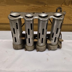 May include: A vintage silver-toned coin dispenser with four cylindrical compartments. Each compartment has a slotted design and a curved top. The dispenser is made of metal and sits on a white surface.