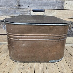 May include: A large, oval-shaped copper container with a matching lid and two side handles. The copper surface shows a weathered, metallic patina with horizontal ridges along the body. A black handle is centred on the top of the flat lid.