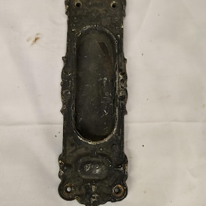May include: An antique bronze-toned recessed door pull with ornate detailing. The rectangular plate features a central oval indentation for gripping, flanked by decorative flourishes and mounting holes. The finish shows signs of age and wear.