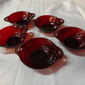 May include: Five ruby red glass bowls with handles. The bowls have a textured design and are oval-shaped. The handles are small and rounded. The bowls are arranged on a white surface.