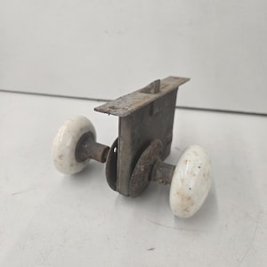 May include: A vintage mortise door lock mechanism featuring two round, off-white porcelain knobs attached to a central metal housing. The metal components show signs of oxidation and wear, with a rectangular faceplate extending from the top.