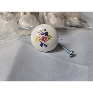 May include: A round white ceramic cabinet knob featuring a colorful floral pattern with pink, blue, and yellow flowers. The knob includes a silver-toned metal screw for installation, and additional knobs are visible in the background in plastic.