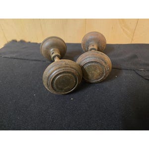 May include: Two round, weathered brass door knobs with a dark, tarnished patina. Each knob features a central circular indentation surrounded by a raised ring, connected to a short metal spindle shaft extending from the back of the hardware.