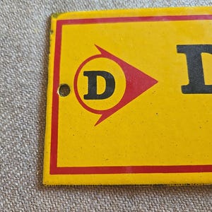 K&ouml;nnte beinhalten: Gelb-rotes Vintage-Metallschild mit dem Buchstaben "D" in einem roten Pfeillogo. Das Schild hat einen roten Rand und ein Loch zum Aufh&auml;ngen. Der Hintergrund ist eine strukturierte, neutrale Farbe.