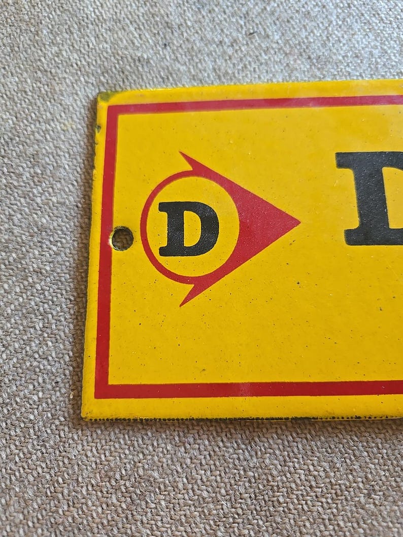 K&ouml;nnte beinhalten: Gelb-rotes Vintage-Metallschild mit dem Buchstaben "D" in einem roten Pfeillogo. Das Schild hat einen roten Rand und ein Loch zum Aufh&auml;ngen. Der Hintergrund ist eine strukturierte, neutrale Farbe.