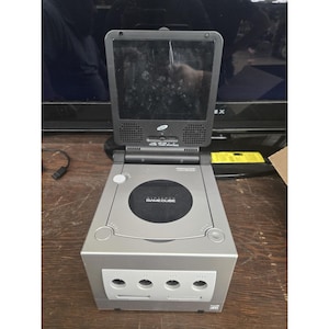 May include: A silver Nintendo GameCube video game console with a black circular disc cover and a gray front panel featuring four controller ports. An attached dark gray flip-up LCD screen is mounted on top, displaying the GameCube logo and text.