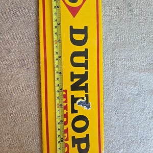 K&ouml;nnte beinhalten: Vintage Dunlop Werbeschild. Das rechteckige Schild ist gelb mit schwarzem Text und einem roten Rand. Das Schild ist etwa 30 cm lang. Das Dunlop-Logo befindet sich oben, darunter das Wort "DUNLOP".