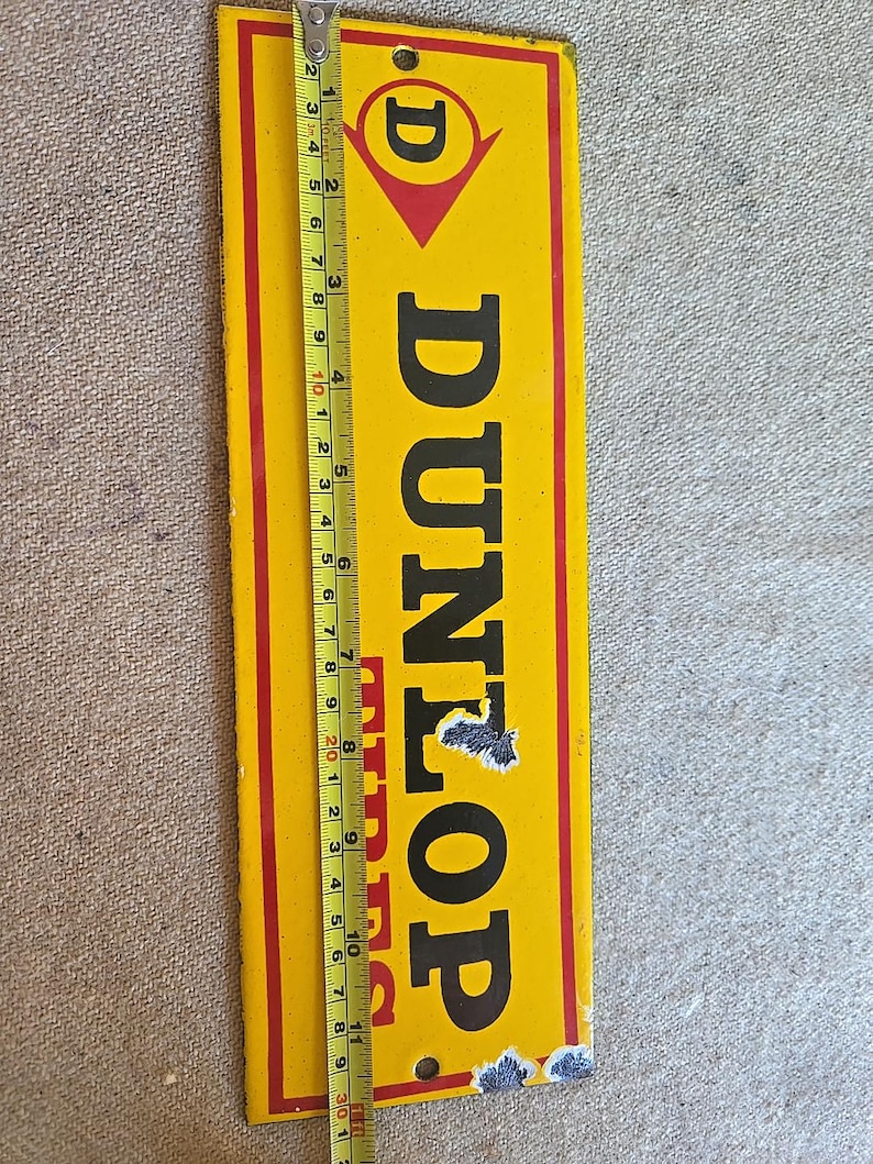 K&ouml;nnte beinhalten: Vintage Dunlop Werbeschild. Das rechteckige Schild ist gelb mit schwarzem Text und einem roten Rand. Das Schild ist etwa 30 cm lang. Das Dunlop-Logo befindet sich oben, darunter das Wort "DUNLOP".