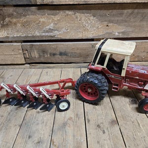 May include: A red and white toy tractor with a plow. The tractor has a beige cab and black tires with red rims. The plow is red with black blades and white accents. The toys are on a wooden surface.